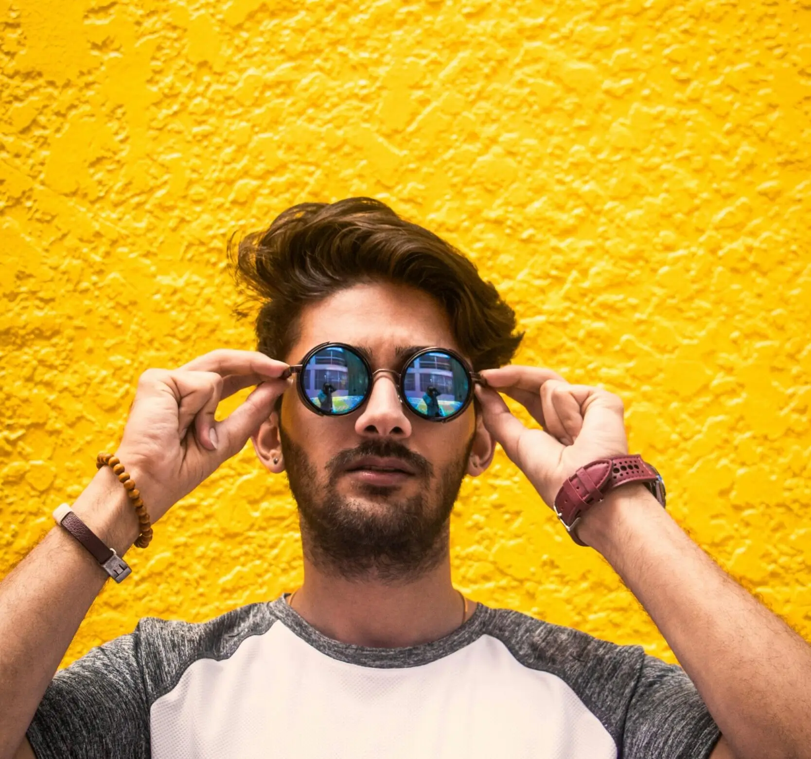 Young man with sunglasses standing against a vibrant yellow wall in daylight.