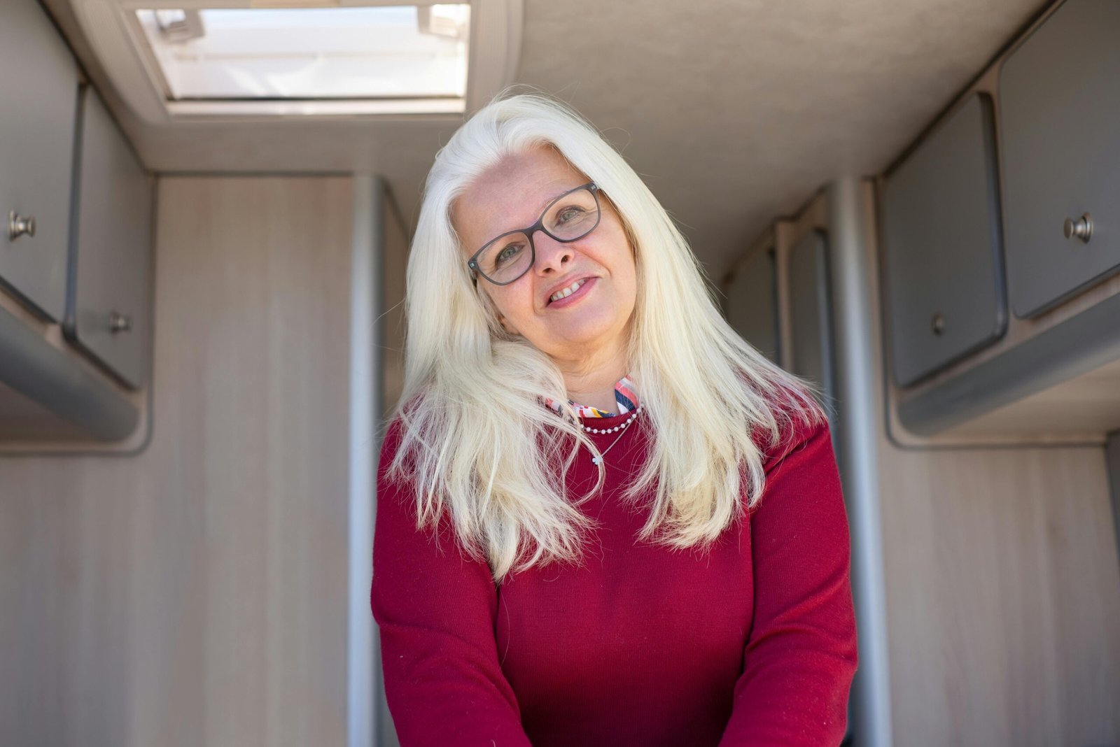 A cheerful woman with glasses and white hair smiling warmly inside a cozy camper van.
